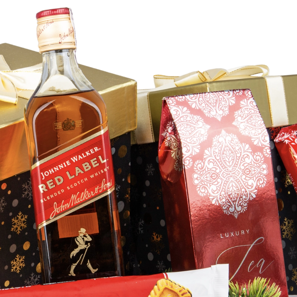 Butelka whisky Johnnie Walker Red Label stojąca na tle świątecznych pudełek prezentowych w czarno-złoty wzór. Obok widoczne jest czerwone opakowanie z napisem "Luxury Tea".