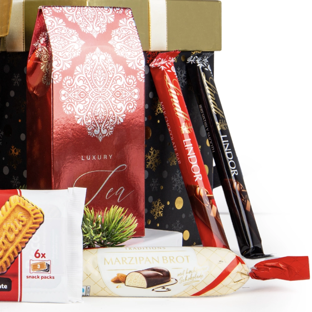 Elementy kosza prezentowego: czerwone opakowanie "Luxury Tea", marcepanowy baton "Marzipan Brot" w czekoladzie, czekoladowe batony Lindor (Milk Chocolate Bar i truffles) oraz opakowanie ciastek. W tle widać świąteczne pudełka.