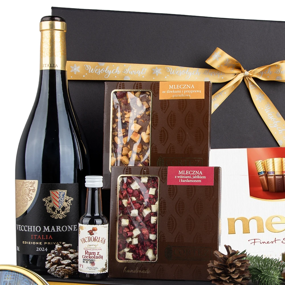 Górna część świątecznego kosza prezentowego z butelką czerwonego wina Vecchio Marone Italia Edizione Private 2024, dwiema tabliczkami ręcznie robionej mlecznej czekolady (jedna ze śliwkami i piernikową przyprawą, druga z wiśniami, jabłkiem i kardamonem), oraz małą butelką likieru Victoria's Rum z czekoladą. W tle widoczna część dużego, czarnego pudełka przewiązanego złotą wstążką z napisem "Wesołych Świąt".