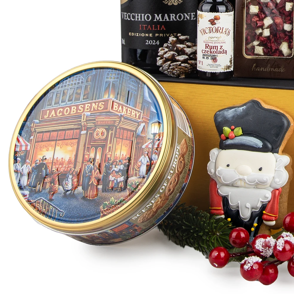 Zbliżenie na elementy świątecznego kosza prezentowego. Na pierwszym planie okrągła, metalowa puszka Jacobsens Bakery z ilustracją ulicznej sceny. Obok niej świąteczny pierniczek w kształcie dziadka do orzechów, ozdobna gałązka igliwia oraz sztuczne czerwone jagody. W tle widoczna część butelki wina Vecchio Marone, mała butelka likieru Victoria's Rum z czekoladą oraz kawałek czekolady z suszonymi owocami.