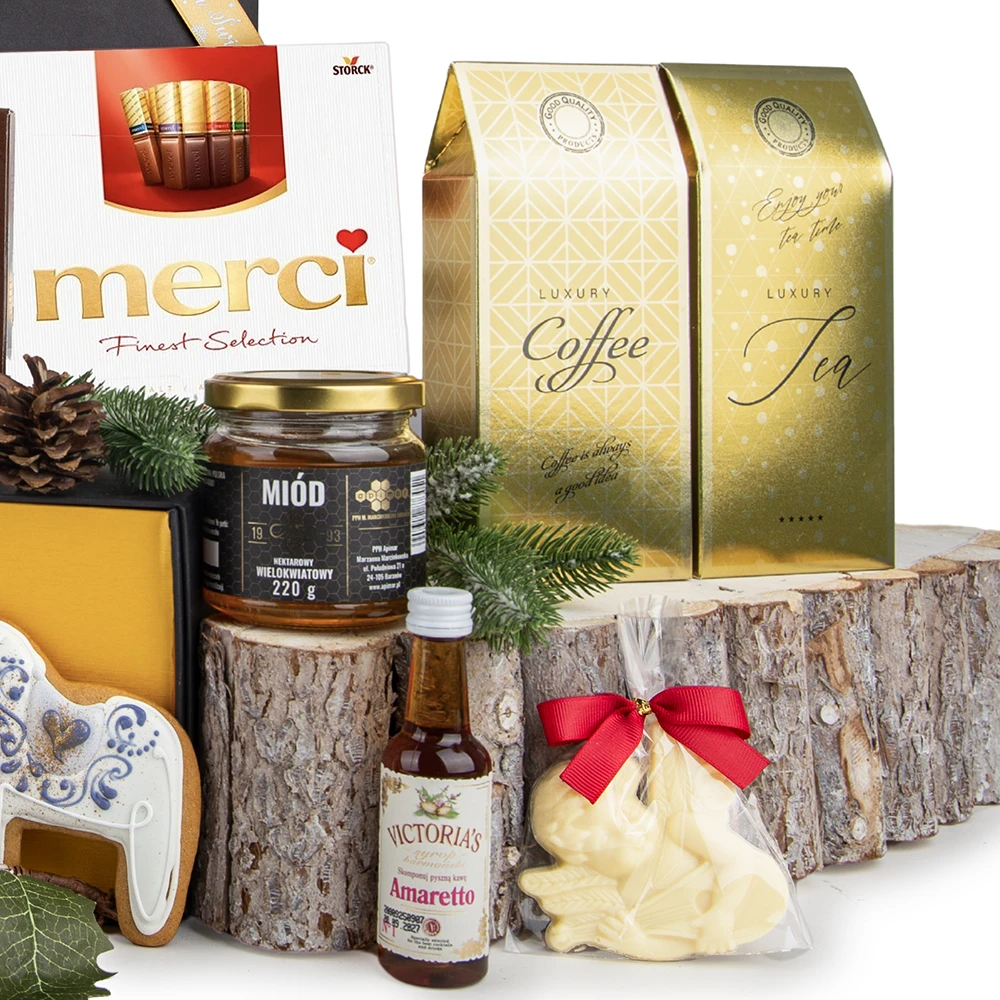 Asortyment słodkości i napojów z kosza prezentowego, ustawiony na drewnianych plastrach. Widoczne są: słoik miodu wielokwiatowego, mała butelka likieru Victoria's Amaretto, czekoladowy królik w torebce z czerwoną kokardą, bombonierka czekoladek Merci, złote opakowania "Luxury Coffee" i "Luxury Tea" oraz świąteczny piernik w kształcie białego konika. Całość udekorowana igliwiem i szyszkami.