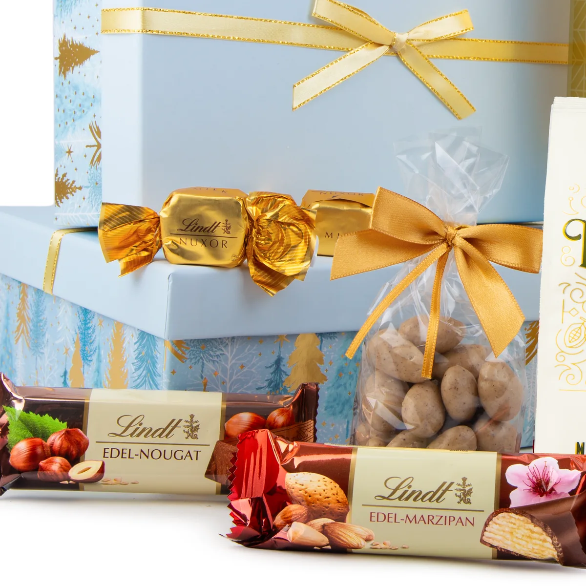Zbliżenie na świąteczny zestaw prezentowy w jasnoniebieskich pudełkach ze złotymi wstążkami, zawierający batony Lindt Edel-Nougat (z orzechami laskowymi) i Lindt Edel-Marzipan (z migdałami), a także złotą czekoladkę Lindt Nuxor oraz woreczek migdałów w polewie.