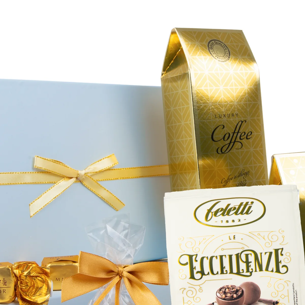 Zbliżenie na jasnoniebieskie pudełko prezentowe przewiązane złotą wstążką, obok złote opakowanie kawy Luxury Coffee i biała torebka pralin Feletti Le Eccellenze.